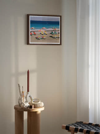 Gerahmter Stranddruck mit bunten Liegen und Sonnenschirmen an beiger Wand