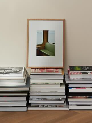 Træindrammet kunsttryk med grøn bænk ved vindue, placeret på en stak bøger mod beige væg