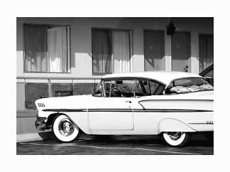 Black-and-white photo of a white vintage car parked outside a motel with curtained windows.