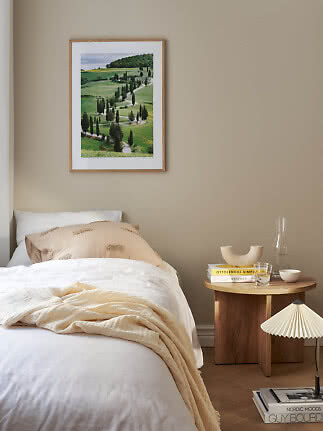 Slaapkamer met bed, ronde houten tafel en ingelijste landschapsprint aan beige muur