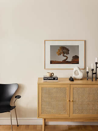 Light wood cabinet with cane doors, candlesticks and framed tree photo against beige wall