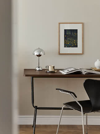 Skrivebord i mørkt træ med kromlampe, kop, åben bog og sort stol ved beige væg