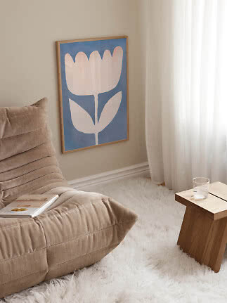 Indrammet blå plakat med abstrakt blomst på væg ved beige loungestol og lille træbord