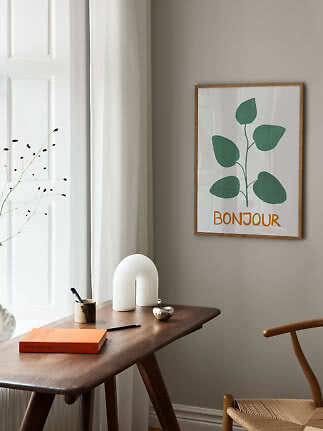 Poster incorniciato con pianta verde e scritta "Bonjour" su parete grigia sopra un tavolo in legno.