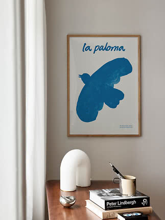 Inramad poster med blå duva på beige vägg ovanför träbord med böcker och kopp