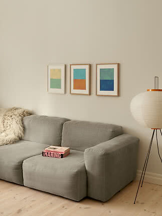Wohnzimmer mit grauem Sofa, drei gerahmten Farbprints an der Wand und weißer Stehlampe.