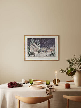 Spisebord med lys, grønt glas og vase under indrammet vintermotiv på beige væg