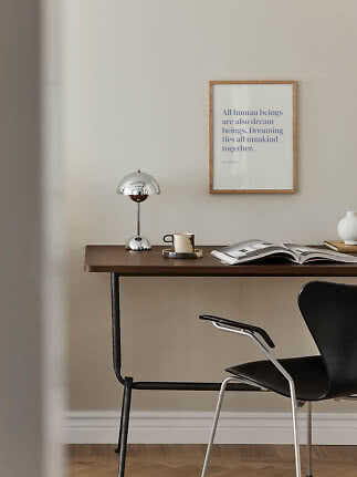 Minimalistisk skrivebord med kromlampe, kop, åben bog og sort stol ved væg med indrammet plakat