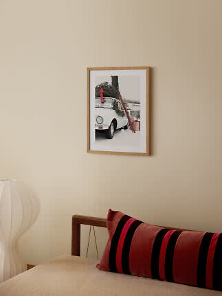 Poster con cornice in legno su parete beige sopra un letto con cuscino rosso a righe.