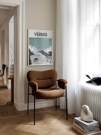 Brun polstret stol med sorte ben ved vindue og Verbier-plakat på beige væg