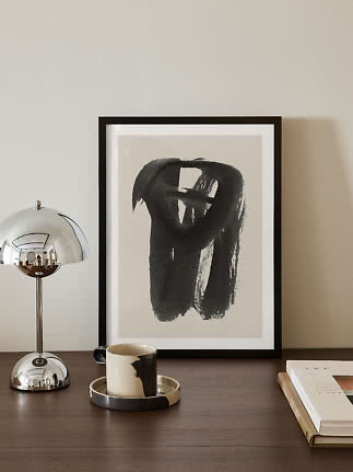 Indrammet abstrakt sort-hvid kunst på træbord med kromlampe, krus og bog