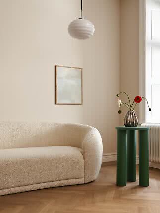 Grønt rundt sidebord med tre ben ved beige sofa, vase med blomster og vindue.