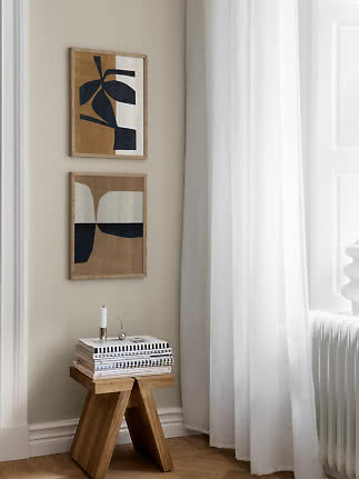 Two abstract framed prints above a wooden stool with magazines beside a bright window