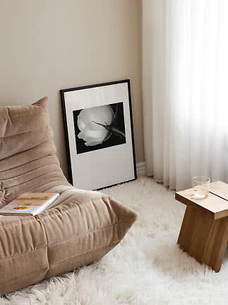 Hyggeligt hjørne med beige lænestol, træbord, hvidt tæppe og indrammet blomsterplakat ved gardin