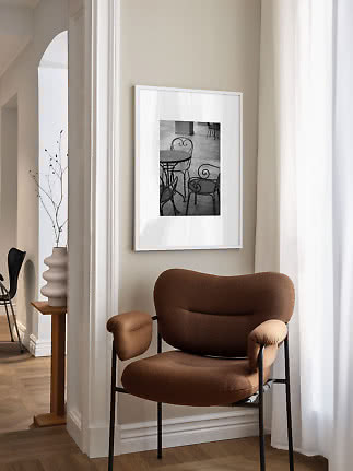 Sillón tapizado marrón junto a pared beige con foto en blanco y negro enmarcada y cortina blanca