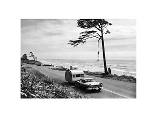 Black-and-white coastal scene with vintage car and trailer on a road beside the ocean