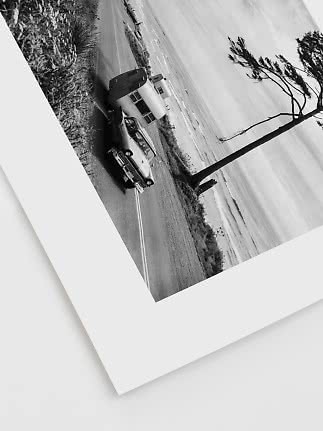 Black-and-white photo of a vintage car towing a caravan on a coastal road by a leaning tree