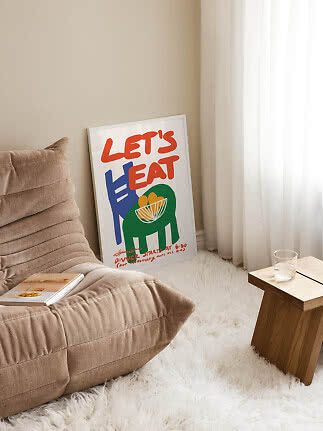 Kleurrijke 'Let's Eat'-poster leunt tegen de muur naast fauteuil, vloerkleed en houten tafeltje