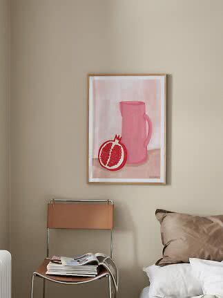Inramad poster med rosa kanna och granatäpple på beige vägg ovanför stol vid säng