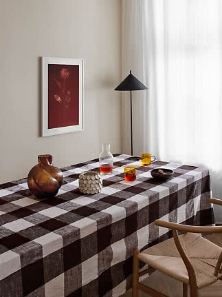 Mesa de comedor con mantel a cuadros marrones, jarrones de vidrio, tazas y lámpara de pie negra