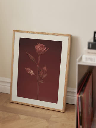 Póster floral enmarcado con una rosa sobre fondo rojo oscuro, apoyado en la pared