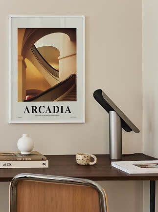 Indrammet Arcadia-plakat med trappemotiv over et skrivebord med lampe, bøger, vase og kop