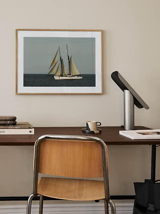 Indrammet billede af en sejlbåd over et skrivebord med lampe, kop og stol