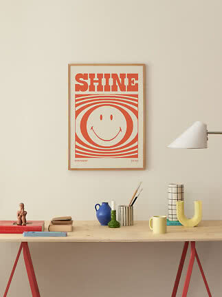Poster incorniciato "SHINE" con smile arancione sopra una scrivania con oggetti decorativi