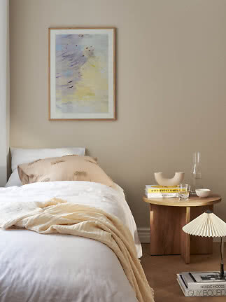 Dormitorio sereno con cama blanca, manta beige, mesa redonda de madera y arte abstracto en la pared.