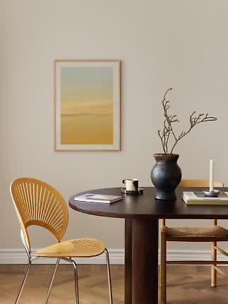Coin repas minimaliste avec table ronde foncée, chaise jaune, vase avec branches et affiche encadrée