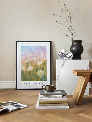 Indrammet blomstermotiv lænet mod væg ved bøger, kop og sort vase med grene.