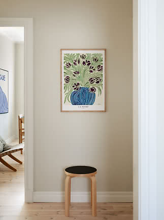 Indrammet blomsterplakat med lilla tulipaner i blå vase over en lys træskammel på beige væg