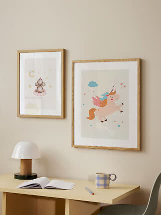 Two framed children's prints with a mouse and unicorn above a desk with lamp, book and mug.