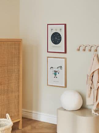 To indrammede børneplakater på beige væg over en bænk med rund hvid lampe og knagerække.