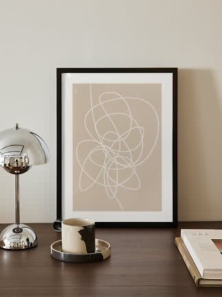 Framed abstract print with white line art on beige background on a dark table with lamp and mug
