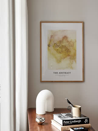 Indrammet abstrakt plakat i gule toner over et træbord med bøger og dekorationer