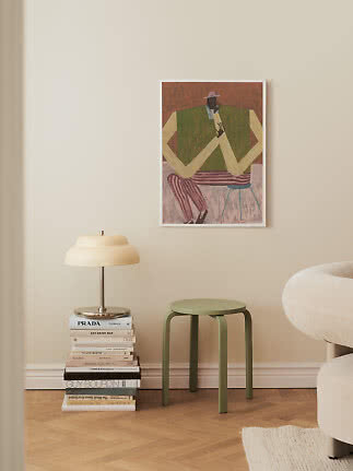 Taburete verde junto a pila de libros y lámpara de mesa bajo cuadro en pared beige