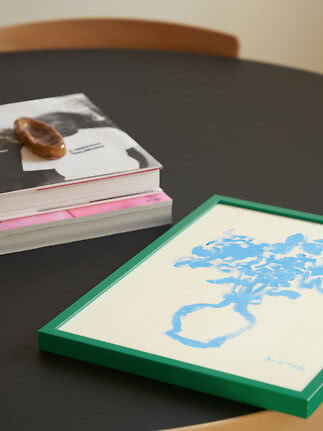 Green framed print with blue vase illustration on a black table beside stacked books