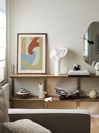 Living room with wall shelf, framed abstract print, white table lamp, books, vases and mirror.