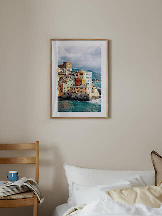 Framed coastal print with colorful houses above the sea on a beige wall over a bed