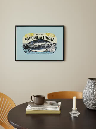 Indrammet plakat med sardindåsemotiv i blå og gul over et mørkt bord med bøger, krus og lysestage