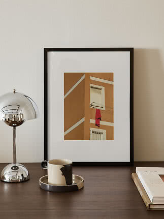 Framed print of an ochre building with a red garment on a clothesline, displayed on a dark table