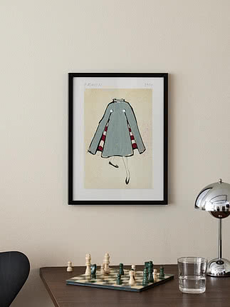 Gerahmter Modeprint mit Skizze eines hellblauen Umhangs an beiger Wand über einem Tisch mit Schachbrett