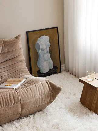 Framed painting of a torso leaning against the wall beside a lounge chair and sheer curtains