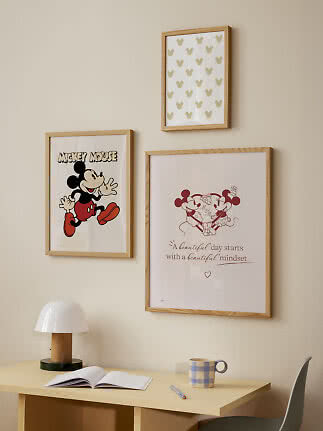 Drei gerahmte Mickey-Mouse-Poster über einem hellen Schreibtisch mit Lampe, Notizbuch und Becher.