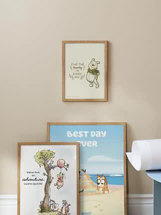 Drei gerahmte Kinderposter mit Holzrahmen an beiger Wand, eins hängt, zwei lehnen auf dem Boden.
