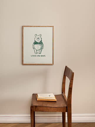 Poster incorniciato di Winnie the Pooh su parete beige sopra una sedia in legno con libri