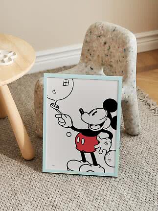 Ingelijste Mickey Mouse-poster met lichtblauwe lijst, leunend tegen een kleine stoel op een vloerkleed