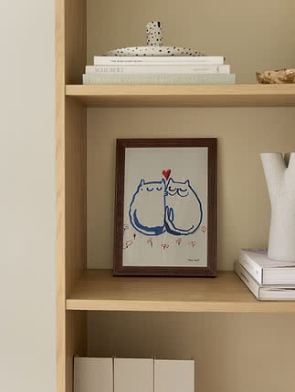 Træreol med indrammet katteillustration, bøger og hvid vase.