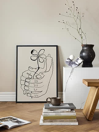 Minimalistisk stuehjørne med indrammet stregtegning, bogstak, kop og mørk vase med tørre grene.